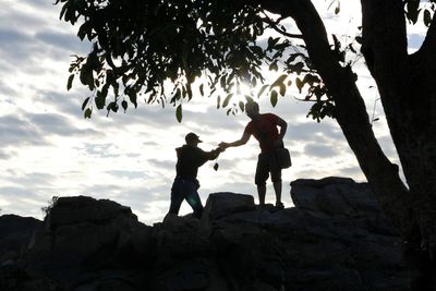 Low angle view of silhouette friends standing on rock against sky