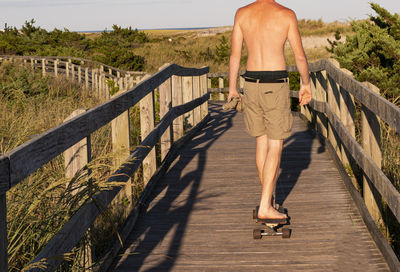 A barefoot man is ridding a skateboard on the fire island lighthouse boardwalk path with no shirt.