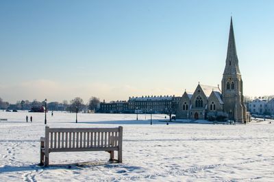 Empty bench on snow covered field with church in background against sky