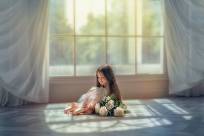 Full length of cute girl with flowers sitting on floor by window at home