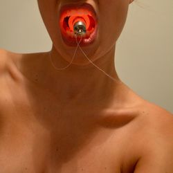 Close-up of shirtless woman holding illuminated bulb in mouth against gray background