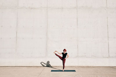 Trendy fit female athlete in sportswear doing extended hand to big toe yoga pose on sports mat while training alone on street against concrete wall in sunny day