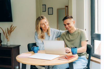 Young happy couple in love sitting home at the table, drinking coffee and working on the laptop.