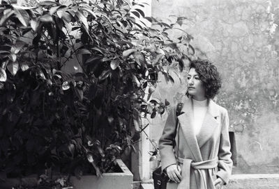 Woman looking away while standing by plants against wall