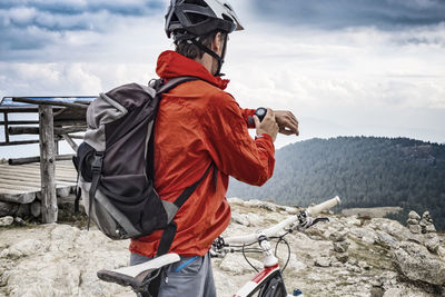 Rear view of mountain biker looking at smart watch against cloudy sky