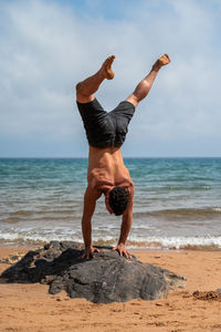 Back view of unrecognizable shirtless male doing adho mukha vriksasana on stone against waving sea and cloudy sky during yoga session on sandy beach in summer