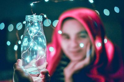 Young woman wrapped in blanket holding jar with illuminated string light at home