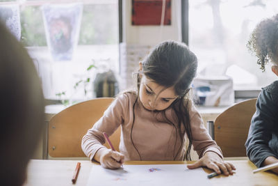 Girl coloring in book with pencil color by female friend at day care center