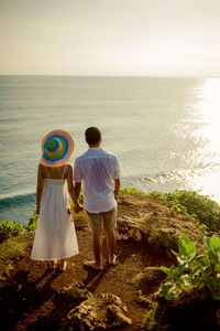 Rear view of couple holding hands while standing on rock at beach against sky during sunset