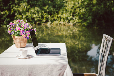 Laptop with coffee and flower on table near the water in the countryside.