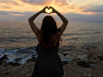 Rear view of woman making heart shape with hands while standing on shore against cloudy sky during sunset