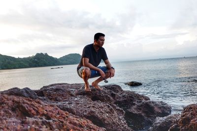 Young man crouching on rock at beach against sky