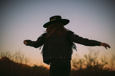 Rear view of woman standing against sky, fringe jacket and hat in the desert