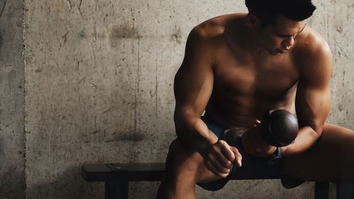 Shirtless man holding dumbbell while sitting in gym
