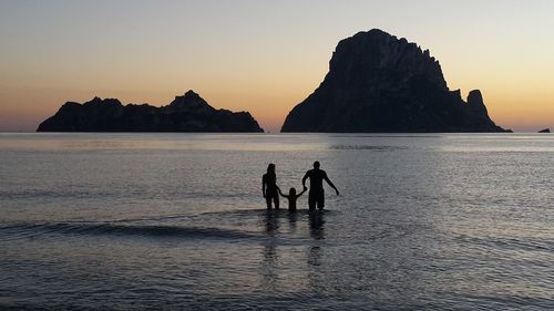 Silhouette people on rocks by sea against sky during sunset