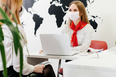 Travel agent having a client in light modern office of tour agency assistant speaking in face mask 