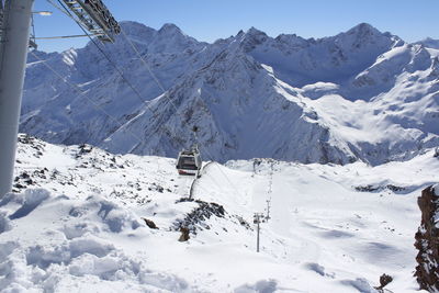Overhead cable car over snow covered field by mountains against clear blue sky