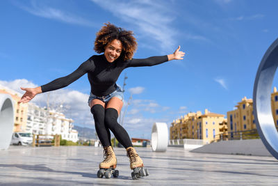 Portrait of mid adult woman with arms outstretched roller skating against blue sky in city