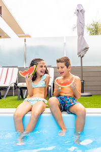 Two funny kids eating watermelon at the the edge of the pool on a summer day