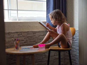 Side view of girl drawing on paper while sitting on chair at home