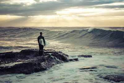 Rear view of man holding surfboard while standing on rock at shore