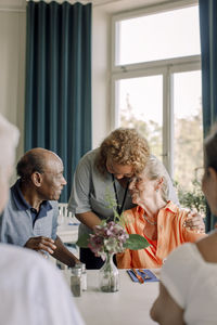Young female caregiver embracing senior woman sitting with friends at dining table in nursing home