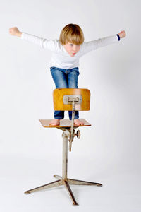 Full length of playful boy with arms outstretched standing on chair against white background