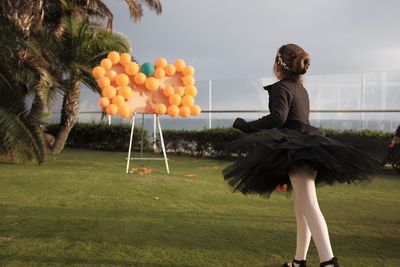 Girl aiming balloons while standing on grassy field against sky