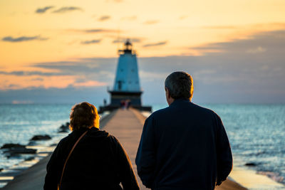Rear view of people walking towards lighthouse amidst sea against sky during sunset