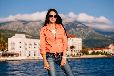 Woman in sunglasses standing at lake against mountains
