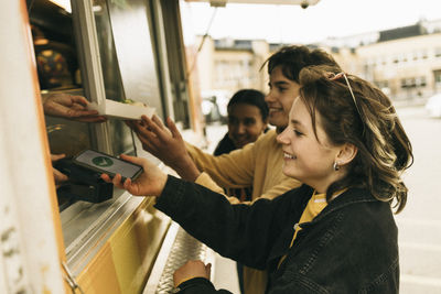 Smiling young woman doing online payment through smart phone while buying food from concession stand