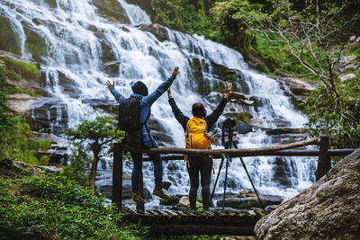 Rear view of friends with arms raised standing by camera and waterfall in forest