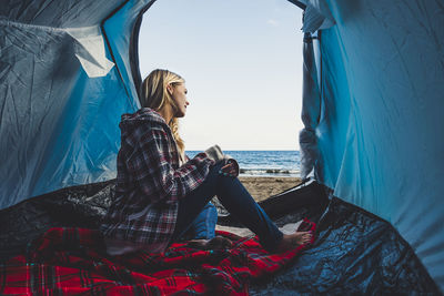 Young woman sitting in tent at beach against sky