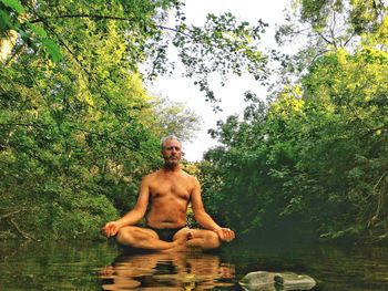 Portrait of shirtless young man by tree against lake