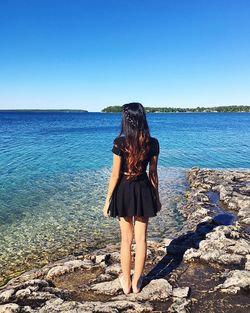 Full length rear view of girl standing in black dress at beach against clear blue sky on sunny day