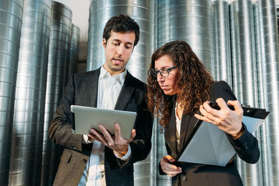 Low angle of smart coworkers in formal suits standing in modern warehouse and discussing business issues while using tablet together