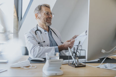 Mature doctor with document smiling at video call on computer in doctor's office
