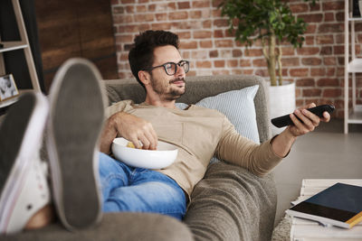 Content man lying on couch at home with bowl of potato chips watching tv