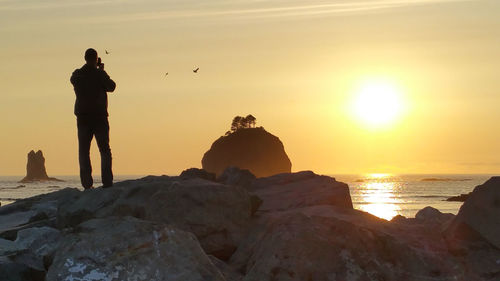 Man standing on rock at beach against sky during sunset