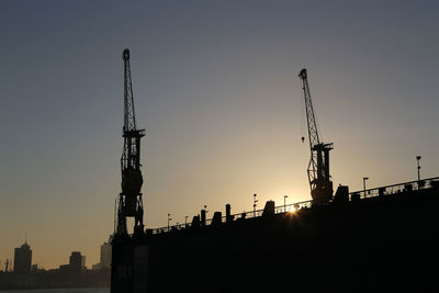 Silhouette cranes at shipyard against sky during sunset