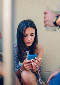 Young woman using mobile phone while sitting against wall