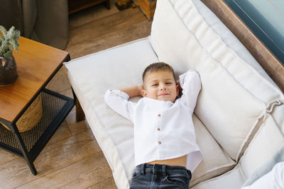 A baby boy is lying on a white sofa on his back, with his hands behind his head. top view