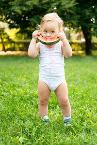 Funny smiling kid boy in white bodysuit eating watermelon at green lawn