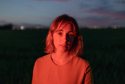 Dreamy young female in vintage style dress looking a camera thoughtfully while standing alone in dark grassy field against cloudy sundown sky in twilight