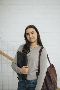 Portrait of confident female student with folder standing against wall in university