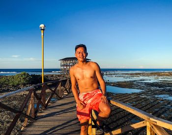 Portrait of shirtless young man standing at beach against sky