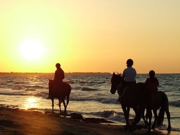 People riding horse on beach against sky during sunset