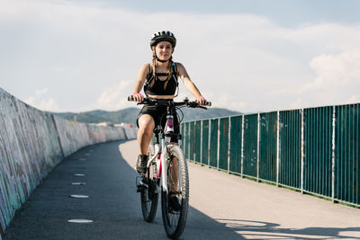 Low angle of confident young female bicyclist in sportswear and helmet riding bike on fenced curvy paved track while training alone in summer day
