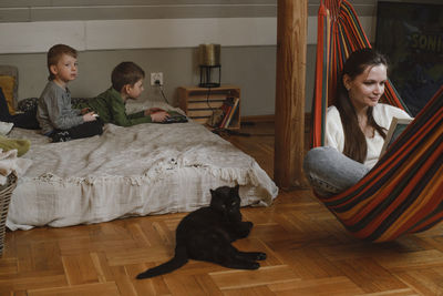 Kids are playing play station while mother is reading book in hammock. family with black cat as a