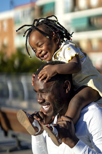 Happy african american girl with dark braids sitting on shoulders of cheerful father and jumping while having fun together on street in sunlight